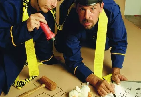 Two actors dressed as police officers humorously investigate a mock crime scene in a playful setup for a Murder Mystery Dinner event. They are kneeling on the ground, surrounded by yellow police line tape, with a chalk outline on the floor resembling the shape of a duck. One officer is holding a red flashlight while the other points at a toy duck with a sign labeled 'Dead Duck.' The scene is lighthearted and comedic, emphasizing the fun, interactive nature of the mystery event.