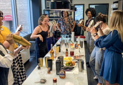 Team shaking cocktail shakers in a mixology class, enjoying the process of creating drinks together. The event promotes collaboration, creativity, and teamwork in a fun and interactive setting.