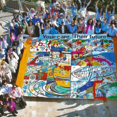 A large group of participants gathers around a completed mural during The Big Picture team building event, celebrating their collaborative artwork with hands raised in excitement. The vibrant mural emphasizes teamwork and shared goals. thumbnail
