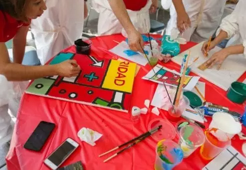 Participants work together on a colorful mural panel during The Big Picture team building event. The table is filled with paint supplies, and one of the participants carefully paints a section featuring bold shapes and colors.