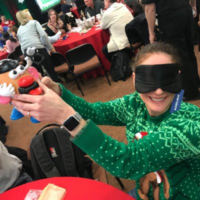 A person wearing a green holiday sweater sits at a table in a large room filled with people. They are holding up a Mr. Potato Head toy while wearing a blindfold, smiling as they participate in an activity. Other attendees in the background are engaged in similar team building holiday-related activities, with festive decorations and red tablecloths creating a cheerful atmosphere. thumbnail