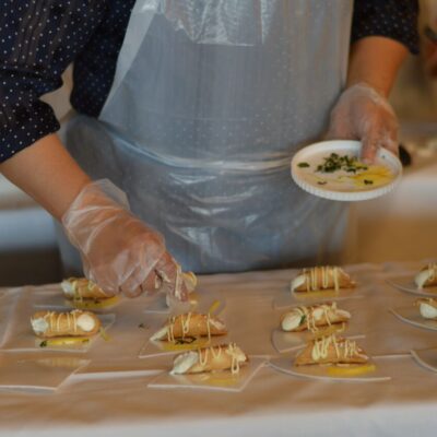 The image shows a participant carefully preparing and plating small tacos as part of a team building food truck challenge. The individual uses gloves for food safety while arranging the tacos neatly on a table covered with a white cloth. Each taco is garnished with a drizzle of sauce, and the participant holds a small plate with additional garnishes, ready to add the finishing touches. thumbnail