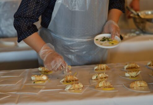 The image shows a participant carefully preparing and plating small tacos as part of a team building food truck challenge. The individual uses gloves for food safety while arranging the tacos neatly on a table covered with a white cloth. Each taco is garnished with a drizzle of sauce, and the participant holds a small plate with additional garnishes, ready to add the finishing touches.