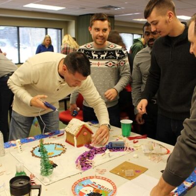 A group of men working together on decorating a gingerbread house during a team building event. One man uses a hot glue gun to assemble pieces, while others discuss and add decorations to the house on a festive, holiday-themed table. thumbnail