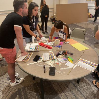 Team members collaborate at a table filled with supplies, working on their bridge design for the Bridging the Divide team building activity. thumbnail