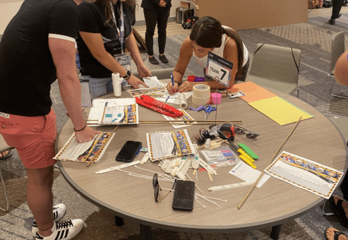Team members collaborate at a table filled with supplies, working on their bridge design for the Bridging the Divide team building activity.