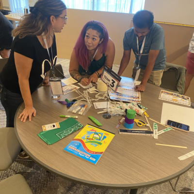 Team 3 members brainstorm and plan their bridge design at a table covered with materials during the Bridging the Divide team building activity. thumbnail