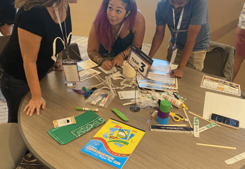 Team 3 members brainstorm and plan their bridge design at a table covered with materials during the Bridging the Divide team building activity.