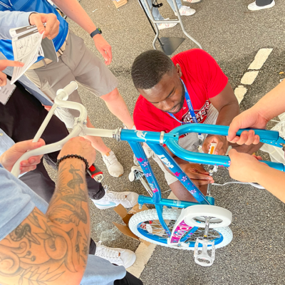 A group of participants working together to assemble a children's bicycle during a Charity Bike Build event. One person is tightening a bolt while others hold the bike steady and refer to instructions, demonstrating teamwork and collaboration. thumbnail