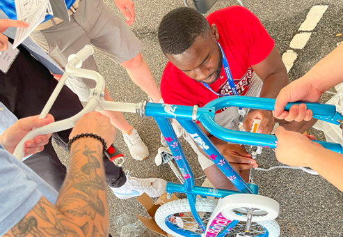 A group of participants working together to assemble a children's bicycle during a Charity Bike Build event. One person is tightening a bolt while others hold the bike steady and refer to instructions, demonstrating teamwork and collaboration.