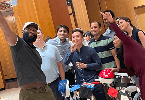 Group of enthusiastic participants taking a selfie during a Charity Bike Build event. They are smiling and posing with peace signs while standing next to the bicycles they assembled, which are adorned with helmets and decorations, highlighting the fun and collaborative atmosphere of the event.