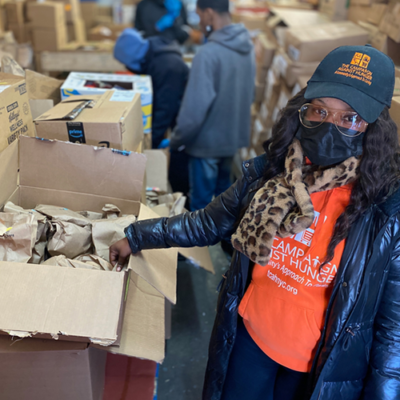 Volunteer wearing a mask and orange shirt organizing brown paper bags in a donation center filled with boxes. thumbnail