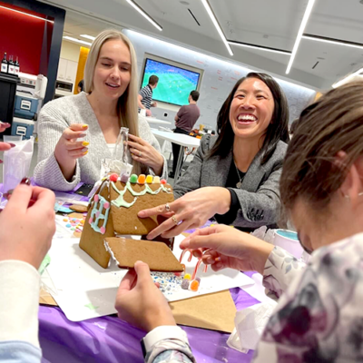 Two women are laughing and enjoying a gingerbread house competition during a team building event. They are actively working together to construct and decorate a small gingerbread house with colorful candies. The setting appears to be a bright, modern office space, and other team members can be seen in the foreground, contributing to the creative project. thumbnail