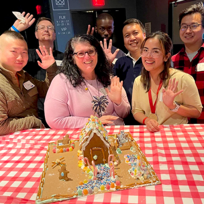 A cheerful group of seven participants standing behind their gingerbread house creation during a team building competition. The gingerbread house is decorated with colorful candies, frosting, and holiday-themed details. The team members are smiling and waving at the camera, showcasing their collaboration and creativity thumbnail