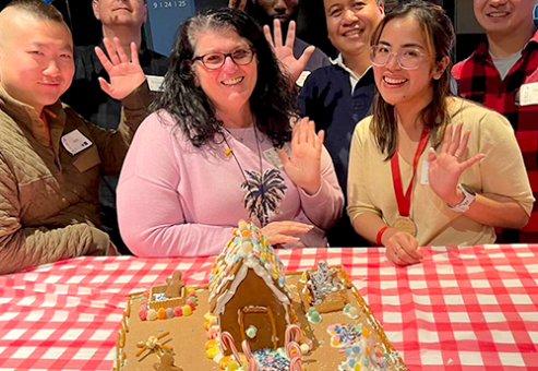 A cheerful group of seven participants standing behind their gingerbread house creation during a team building competition. The gingerbread house is decorated with colorful candies, frosting, and holiday-themed details. The team members are smiling and waving at the camera, showcasing their collaboration and creativity