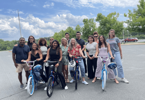 A group of smiling participants standing outdoors with two completed bicycles during a Charity Bike Build event. The team poses together in a parking, showing their assembled bikes with helmets on the handlebars.