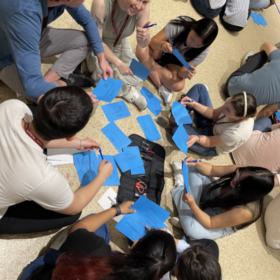 A group of people sitting in a circle on the floor during a team building activity, each holding blue cards with instructions. They are engaging in a collaborative task, exchanging ideas and writing down notes. thumbnail