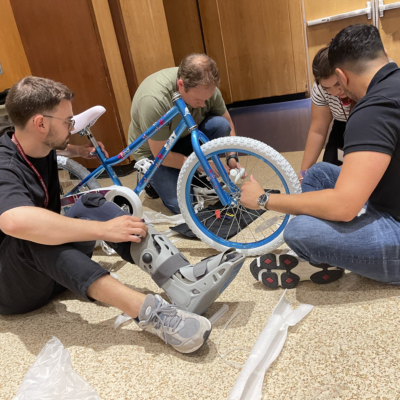 Group of four participants sitting on the floor working together to assemble a blue children's bike during a Charity Bike Build team building event. The participants are focused on attaching parts to the bike frame, demonstrating teamwork and collaboration. thumbnail