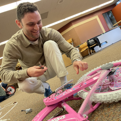 A man crouches on the floor with a smile, carefully assembling a pink bicycle during a Charity Bike Build team building event. He uses tools to attach the final pieces, showcasing hands-on involvement in creating bikes for children. thumbnail